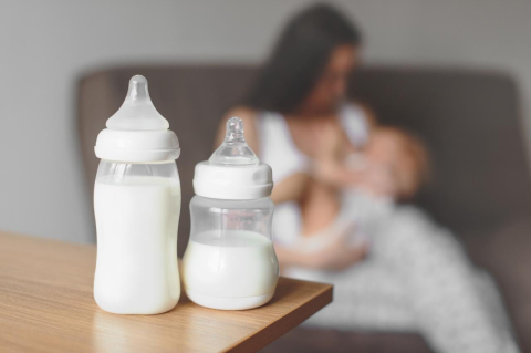 Two bottles of breastmilk on wood table with a mother breast-feeding a child in the background