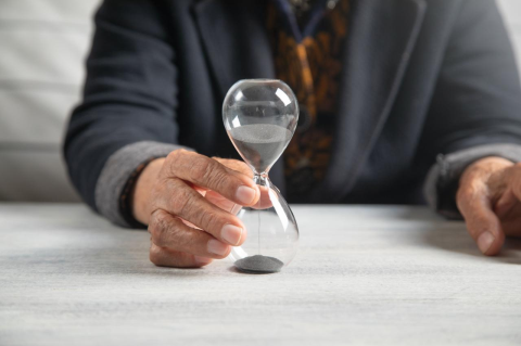a pair of wrinkled hands on a tabletop holds an hourglass with black sand